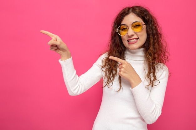 Jonge, mooie vrouw in witte coltrui met een gele bril, blij en vrolijk, wijzend met wijsvingers naar de zijkant die op roze staat