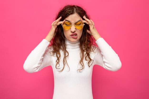 Jonge mooie vrouw in witte coltrui met een gele bril die er niet blij uitziet als ze haar slapen aanraakt met hoofdpijn