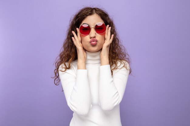 Jonge, mooie vrouw in witte coltrui met een rode bril die er gelukkig en positief uitziet en haar lippen houdt zoals gaan kussen