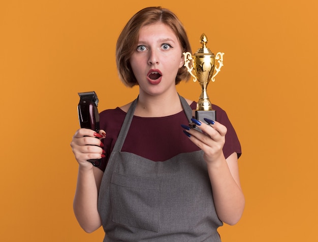 Jonge mooie vrouw kapper in schort bedrijf trimmer machine en gouden trofee kijken voorzijde verrast en verbaasd staande over oranje muur