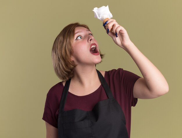 Jonge mooie vrouw kapper in schort houden scheerkwast met scheerschuim kijken verbaasd en verbaasd staande over groene muur