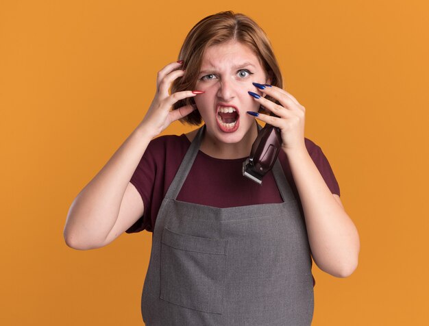 Jonge mooie vrouw kapper in schort met trimmer kijken voorkant schreeuwen met boos gezicht gekke gekke en gefrustreerde staande over oranje muur