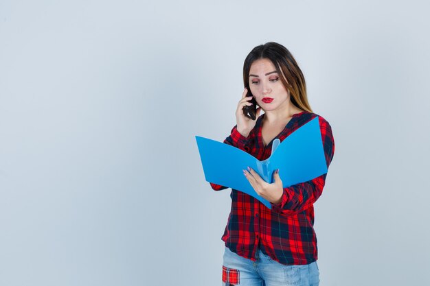 Jonge mooie vrouw kijkt naar de map, praat over de telefoon in een casual shirt, spijkerbroek en kijkt peinzend. vooraanzicht.