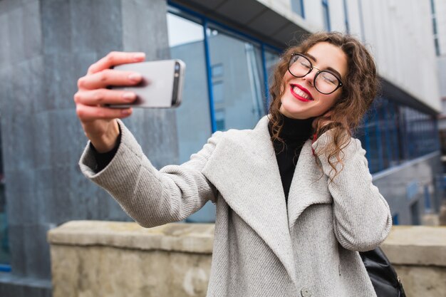 Jonge mooie vrouw lopen in de stad straat in grijze jas herfst fashion stijl, bril, smartphone houden, selfie foto nemen, glimlachen, gelukkig