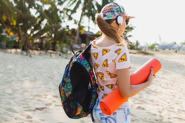 Jonge mooie vrouw lopen op het strand met yogamat, hipster sport swag stijl, denim shorts, t-shirt, rugzak, zonnig, zomerweekend