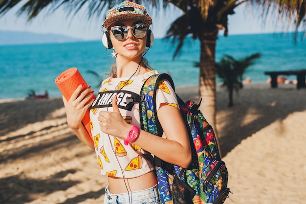 Jonge mooie vrouw lopen op het strand met yogamat, luisteren naar muziek op koptelefoon, hipster sport swag stijl, denim shorts, t-shirt, rugzak, pet, zonnebril, zonnig, zomerweekend, vrolijk