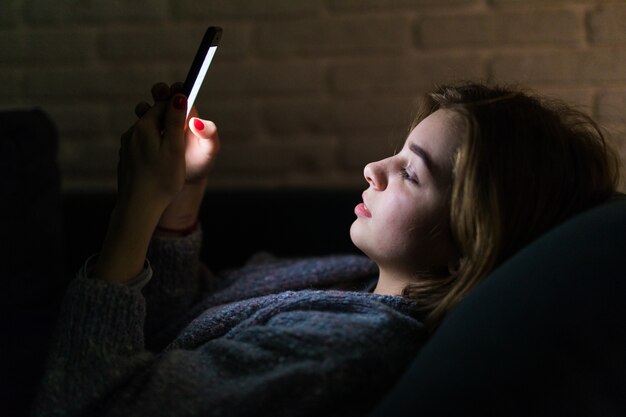 Jonge mooie vrouw met behulp van slimme telefoon 's nachts in bed - technologie, sociaal netwerk, slapeloosheid concept