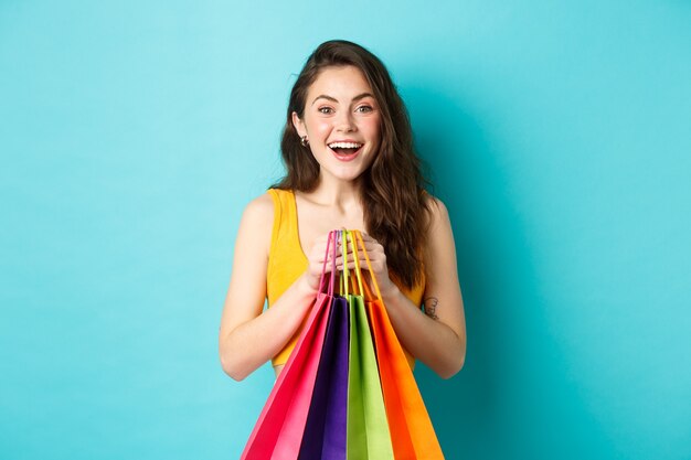 Jonge mooie vrouw met boodschappentassen, glimlachend opgewonden naar de camera, kopend met kortingen, staande over blauwe achtergrond