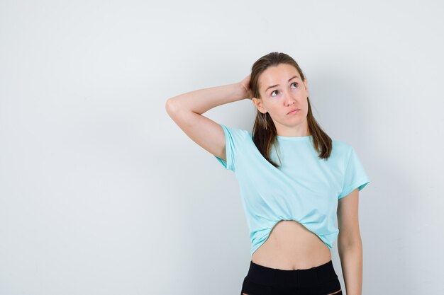 Jonge mooie vrouw met de hand achter het hoofd, wegkijkend in t-shirt en bedachtzaam kijkend. vooraanzicht.