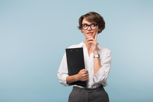 Jonge mooie vrouw met donker kort haar in shirt en bril met map in de hand terwijl ze dromerig opzij kijkt over blauwe achtergrond