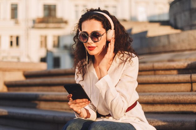 Jonge mooie vrouw met donker krullend haar in zonnebril en witte jas zittend op de trap en het gebruik van mobiele telefoon terwijl ze naar muziek luistert in een koptelefoon op straat