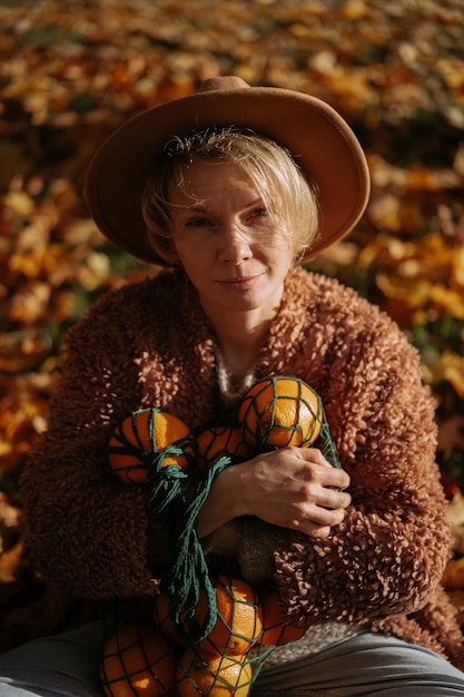 Jonge mooie vrouw met een hoed in een herfstpark, een stringtas met sinaasappels, een vrouw gooit herfstbladeren op. herfststemming, felle kleuren van de natuur.