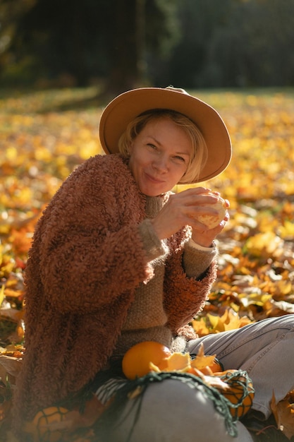 Jonge mooie vrouw met een hoed in een herfstpark, een stringtas met sinaasappels, een vrouw gooit herfstbladeren op. Herfststemming, felle kleuren van de natuur.