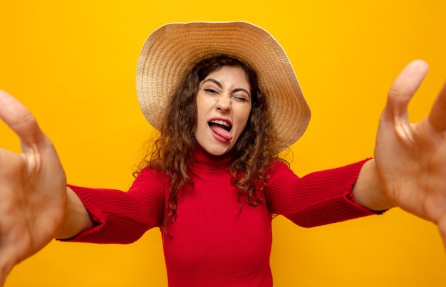 Jonge, mooie vrouw met een rode coltrui in een zomerhoed, blij en vrolijk met plezier terwijl ze haar tong uitsteekt terwijl ze op oranje staat