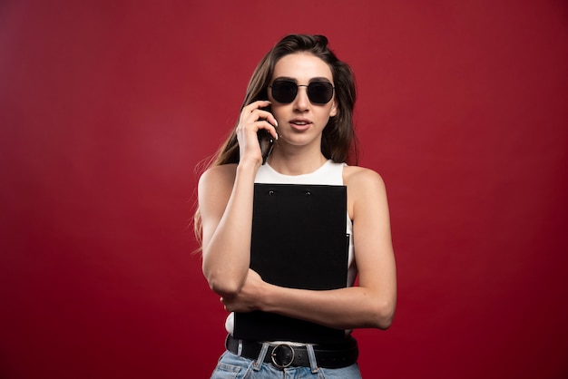 Jonge mooie vrouw met glazen die op telefoon spreken en notitieboekjes houden