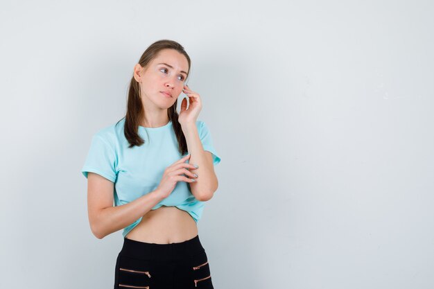 Jonge mooie vrouw met hand in de buurt van gezicht, wegkijkend in t-shirt, broek en peinzend, vooraanzicht.