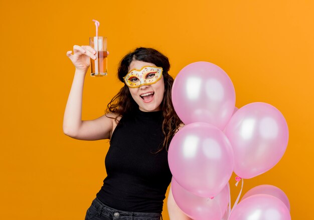 Jonge mooie vrouw met krullend haar bedrijf bos lucht ballonnen en cocktail in partij masker gelukkig en vrolijk verjaardagsfeestje concept permanent over oranje muur