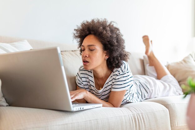 Jonge mooie vrouw met krullend haar die aan laptop computer werken terwijl thuis het zitten op de bank