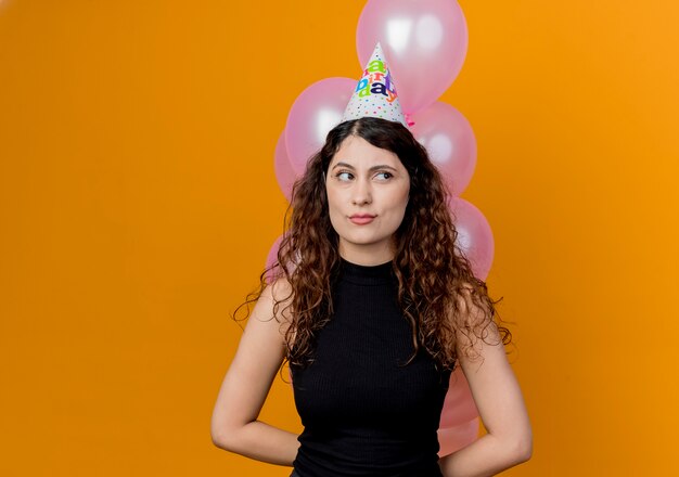 Jonge mooie vrouw met krullend haar die bos van luchtballons houden die opzij kijken met sceptisch expressin concept van de verjaardagsfeestje die zich over oranje muur bevinden