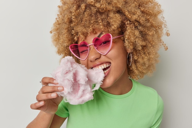 Jonge mooie vrouw met krullend haar eet heerlijke suikerspin geniet van zoete snack draagt roze hart zonnebril en casual groen t-shirt geïsoleerd op witte achtergrond Tienermeisje bijt suikerspin