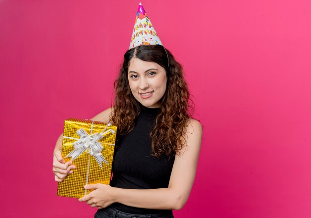 Jonge mooie vrouw met krullend haar in een vakantie GLB bedrijf de doos van de verjaardagsgift die vrolijk het concept van de verjaardagspartij over roze glimlachen