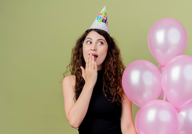 Jonge mooie vrouw met krullend haar in een vakantie GLB die luchtballons houden die opzij verrast en gelukkig over licht kijken