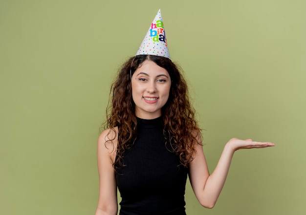 Gratis foto jonge mooie vrouw met krullend haar in een vakantie glb presenteren met arm van hand het glimlachen concept van de verjaardagspartij die zich over oranje muur bevindt