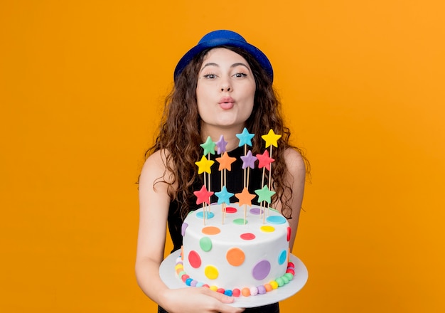 Jonge mooie vrouw met krullend haar in een vakantie hoed met verjaardagstaart blaast een kus blij en positief staande over oranje muur