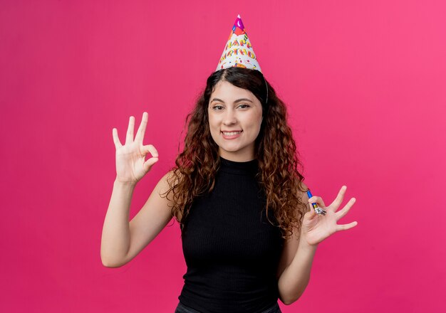 Jonge mooie vrouw met krullend haar in een vakantiepet met fluitje dat ok teken toont die vrolijk het concept van de verjaardagspartij glimlachen die zich over roze muur bevinden