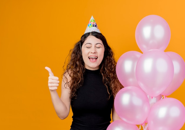 Jonge mooie vrouw met krullend haar in een vakantiepet met luchtballons gek gelukkig duimen opdagen verjaardagsfeestje concept staande over oranje muur