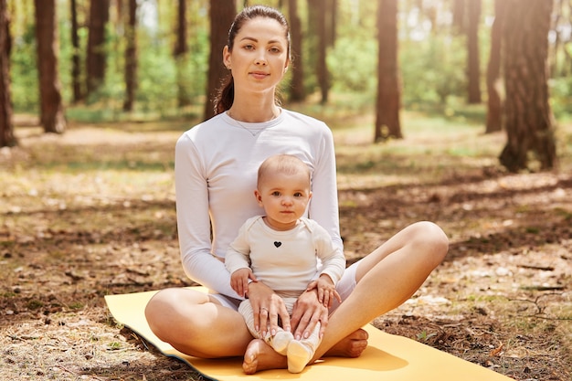 Jonge mooie vrouw met paardenstaart zittend op yogamat met kleine dochtertje en direct naar voren kijkend