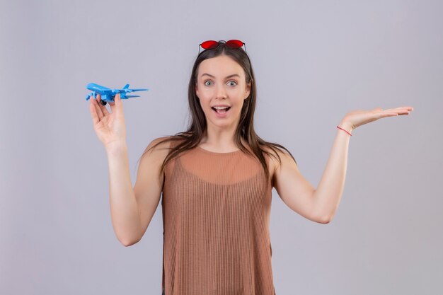 Jonge mooie vrouw met rode zonnebril op hoofd bedrijf speelgoed vliegtuig blij en verrast staan met arm opgeheven op witte achtergrond
