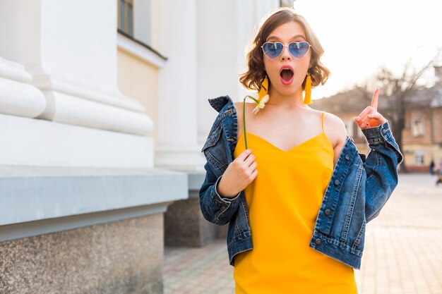 Jonge mooie vrouw met verbaasde gezichtsuitdrukking, emotionele, geschokte emotie, stijlvolle kleding dragen, spijkerjasje, gele top, bloem vasthouden, zonnige zomer, trendy grappige zonnebril