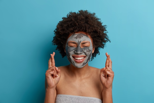 Jonge mooie vrouw past klei voedend masker toe op het gezicht, glimlacht breed, staat in handdoek gewikkeld, kruist vingers, wacht op dromen die uitkomen, staat binnen. Wellness, gezondheidszorg en schoonheidsconcept