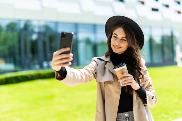 Jonge mooie vrouw student slijtage in jas en hoed koffie te houden om te gaan nemen selfie aan de telefoon in de straat