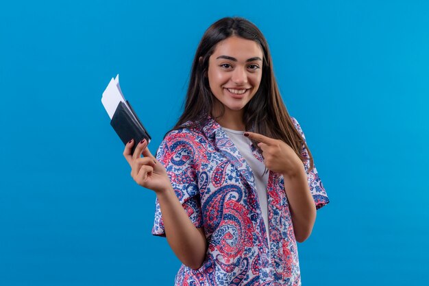 Jonge mooie vrouw toeristische bedrijf paspoort met kaartjes kijken camera wijzend met wijsvinger naar hen glimlachend vrolijk staande over geïsoleerde blauwe achtergrond