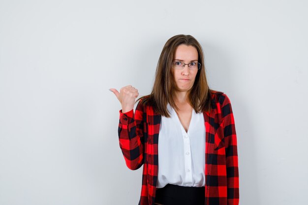 Jonge mooie vrouw wijst naar links met duim in casual outfit en kijkt peinzend, vooraanzicht.