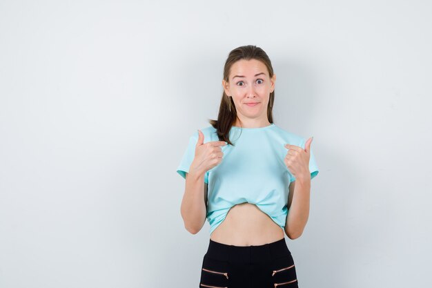 Jonge mooie vrouw wijst naar zichzelf in t-shirt, broek en ziet er vrolijk uit, vooraanzicht.