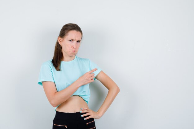 Jonge mooie vrouw wijzend naar de rechterbovenhoek in t-shirt, broek en chagrijnig, vooraanzicht.