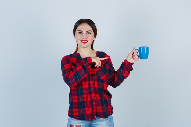 Jonge mooie vrouw wijzend op cup in casual shirt, jeans en vrolijk, vooraanzicht.