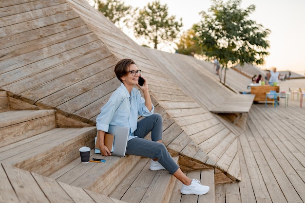 Jonge mooie vrouw zit met laptop in moderne parkstraat met smartphone