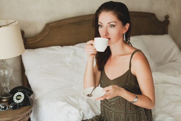 Jonge mooie vrouw zittend op bed in hotel, stijlvolle jurk, sensuele stemming, koffie drinken, beker te houden