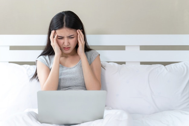Jonge mooie vrouw zittend op bed, ze gebruikt laptop voelen ernstig en hoofdpijn