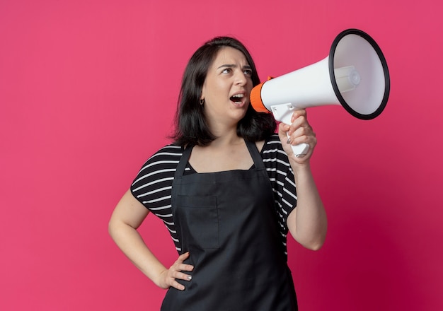 Gratis foto jonge mooie vrouwelijke kapper in schort schreeuwen naar megafoon over roze