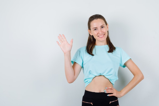 Jonge mooie vrouwelijke zwaaiende hand voor begroeting in t-shirt en op zoek vrolijk, vooraanzicht.