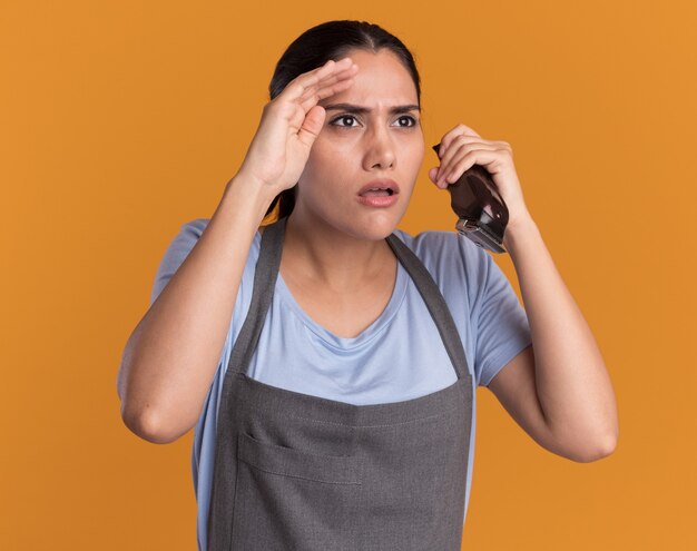 Jonge mooie vrouwenkapper die in de trimmer van de schortholding nauw opzij kijkt met hand boven het hoofd over oranje muur