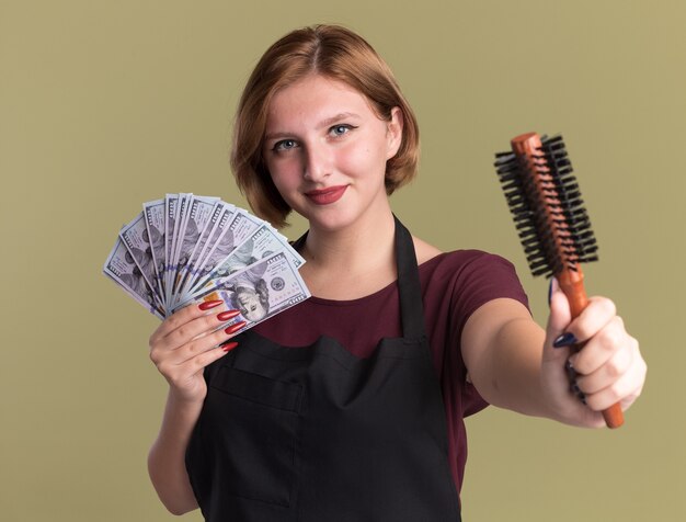 Jonge mooie vrouwenkapper die in het contant geld van de schortholding haarborstel toont die zelfverzekerd over groene muur glimlacht