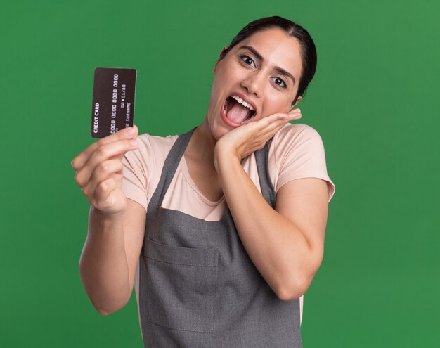 Jonge mooie vrouwenkapper die in schort creditcard het gelukkige en positieve glimlachen toont die zich over groene muur bevindt