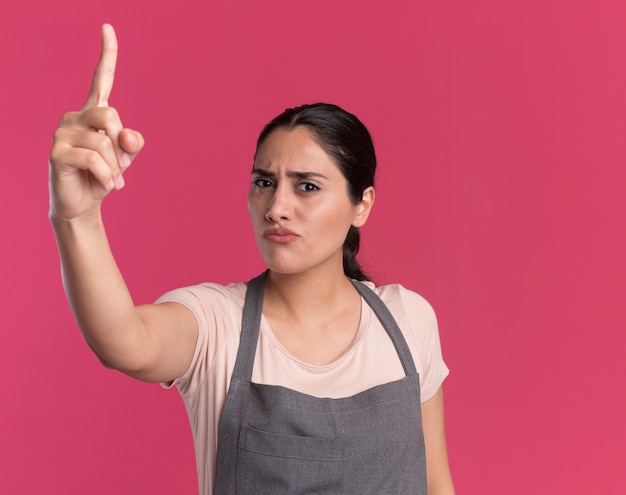 Jonge mooie vrouwenkapper die in schort voorzijde met ernstig gezicht bekijkt dat wijsvingerwaarschuwing toont die zich over roze muur bevindt