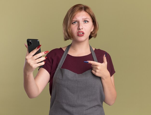 Jonge mooie vrouwenkapper in de smartphone die van de schortholding met wijsvinger richt op het wordt verward status over groene muur